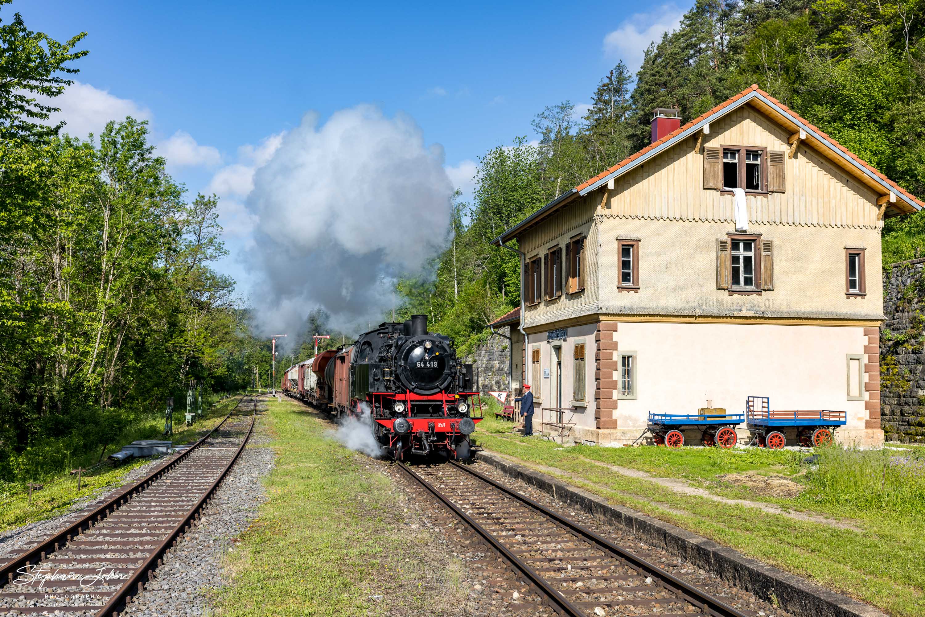 Lok 64 419 erreicht mit einem Güterzug bei der Durchfahrt durch den Bahnhof Grimmelshofen. Mittels K-Scheibe erhält der Lokführer den Auftrag, die Fahrzeit zu kürzen.